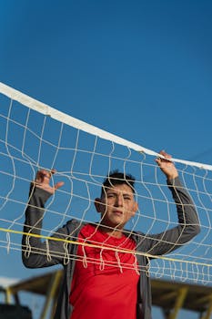 A young man in a red shirt stands outdoors holding a soccer net under a clear blue sky.