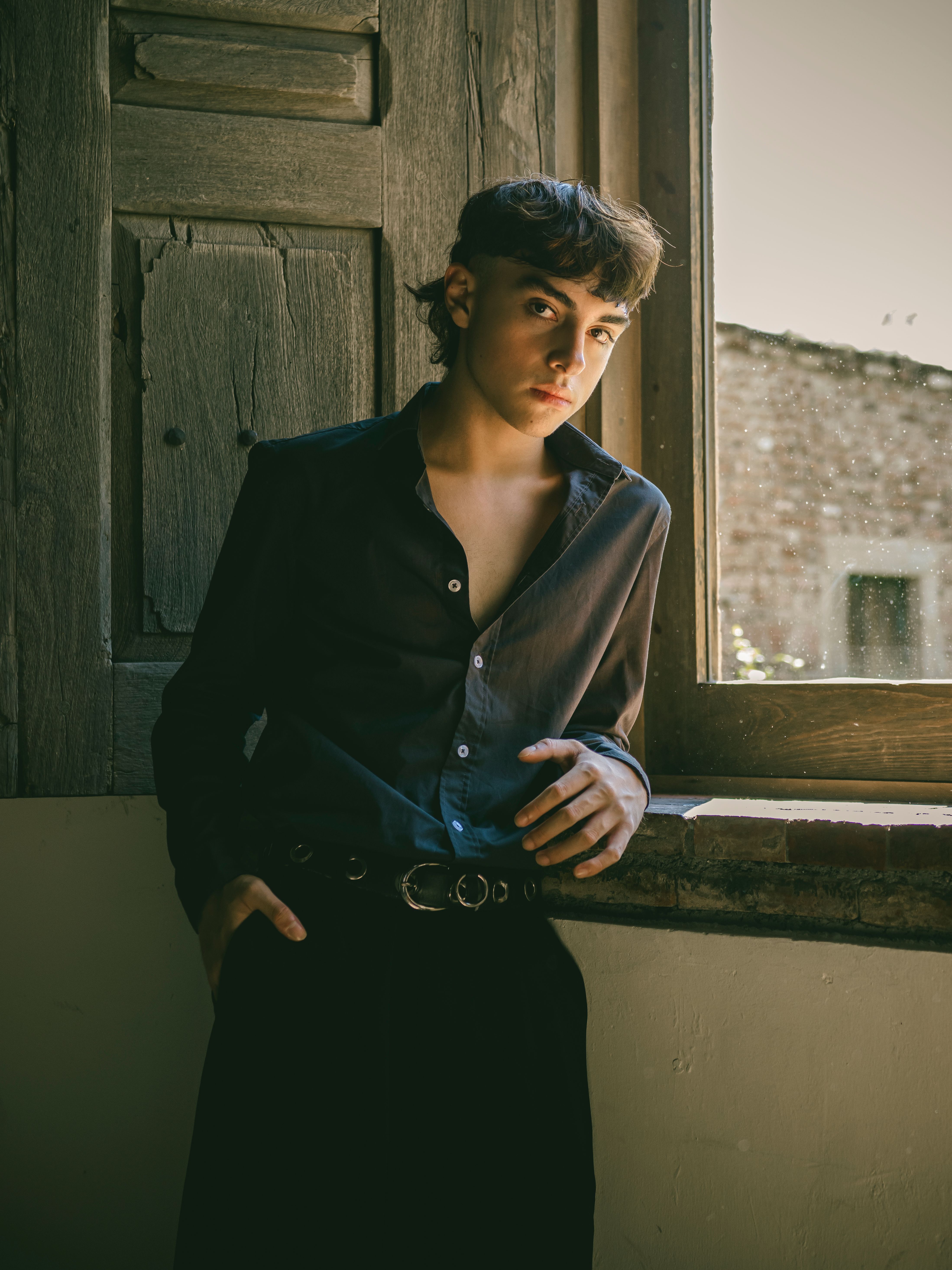 Stylish young adult in a dark shirt gazing through a rustic window indoors, exuding a moody fashion vibe.