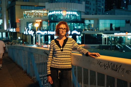 A woman standing on a bridge in a city at night, surrounded by modern buildings.