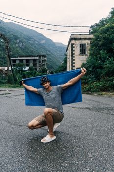 Man with towel poses in a deserted street with mountains and abandoned building in background.
