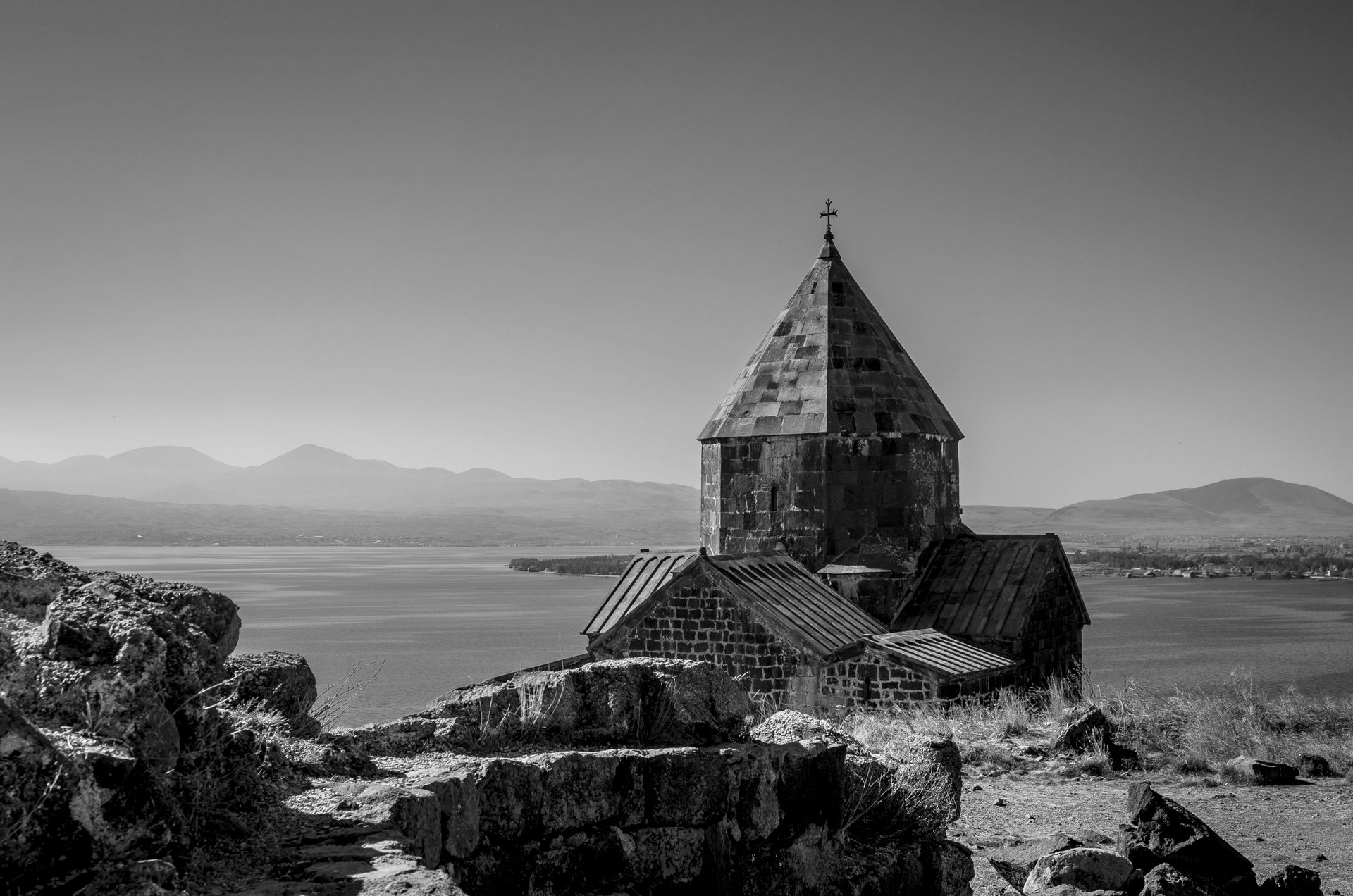 A tranquil monochrome image of a historic chapel by Lake Sevan, Armenia, amid mountains and serene waters.