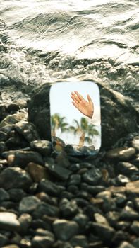 A reflective mirror on a rocky beach with an ocean view and a human hand in the reflection.