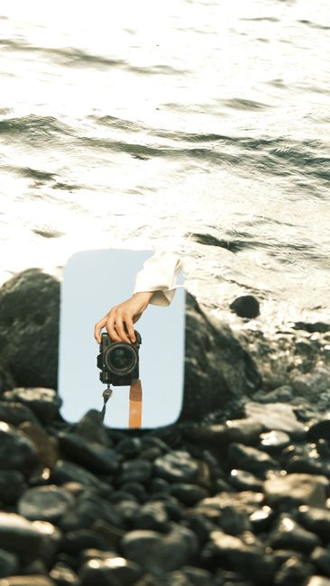 A camera reflection captured on a mirror placed among rocks by the shore, creating an artistic composition.