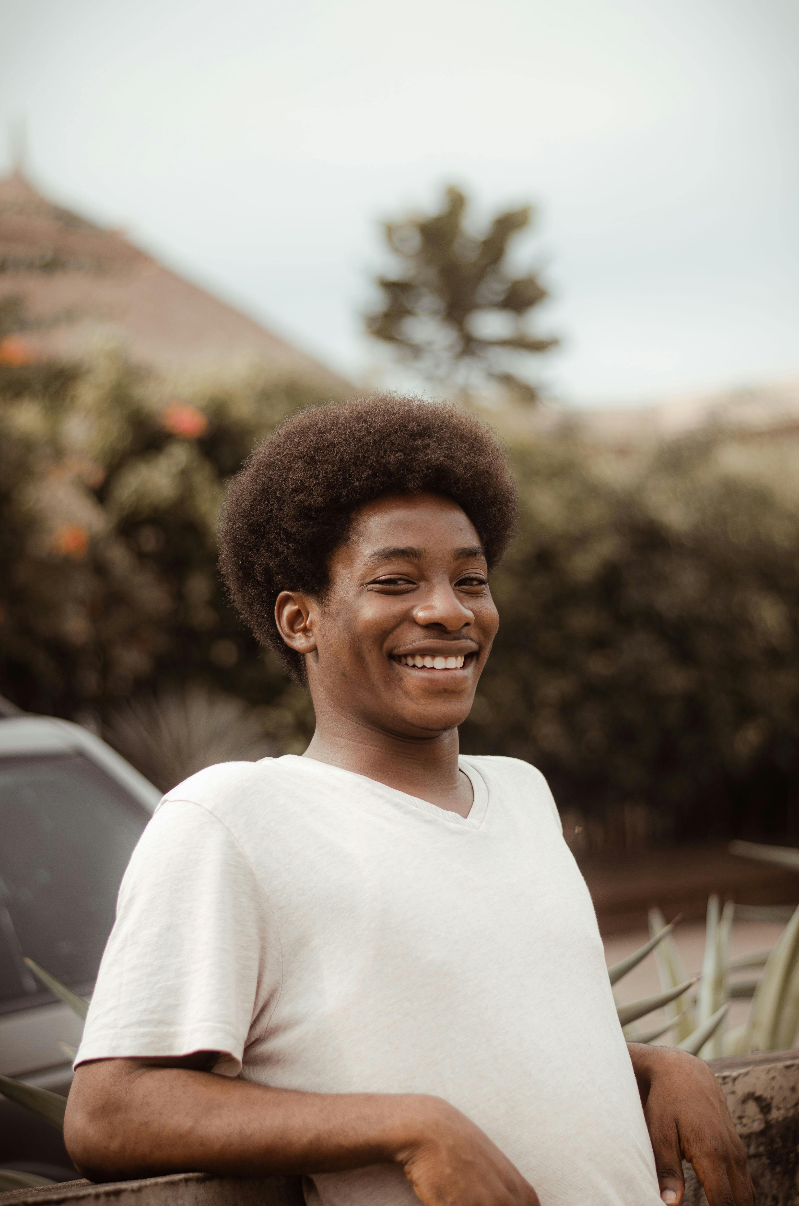A young African man with an afro smiling while leaning against a railing in an outdoor setting.