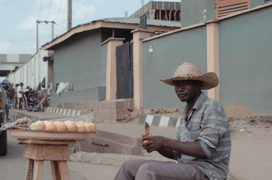 Black Nigerian civil servant man in a formal light-blue shirt standing at an Abuja market stall, carefully inspecting a printed receipt next to bags of rice and beans, natural outdoor daylight, photorealistic