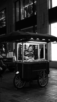Black and white photo of a street vendor selling goods at night under city lights.