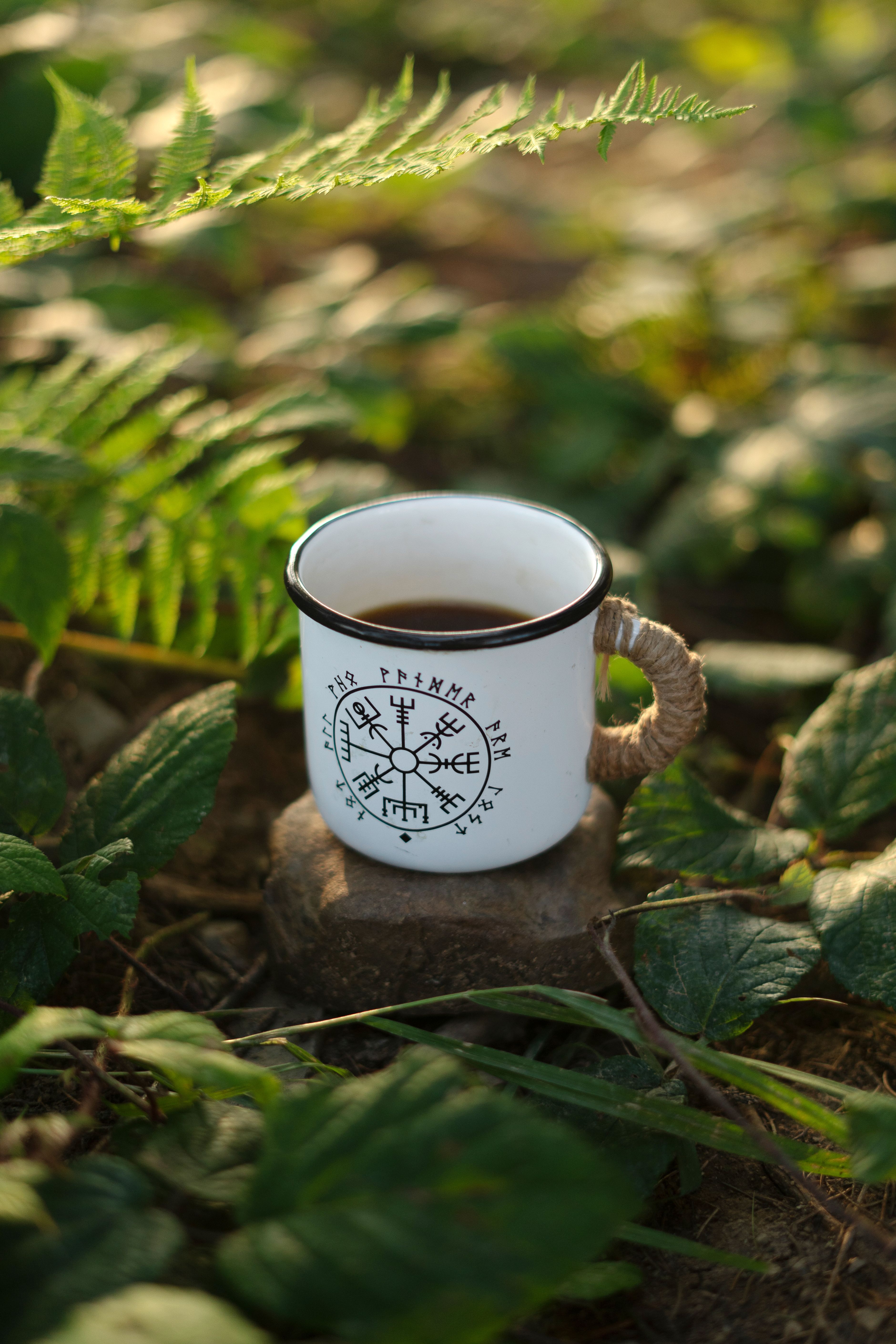 A white enamel mug with a rune design, filled with herbal tea, amidst lush greenery.
