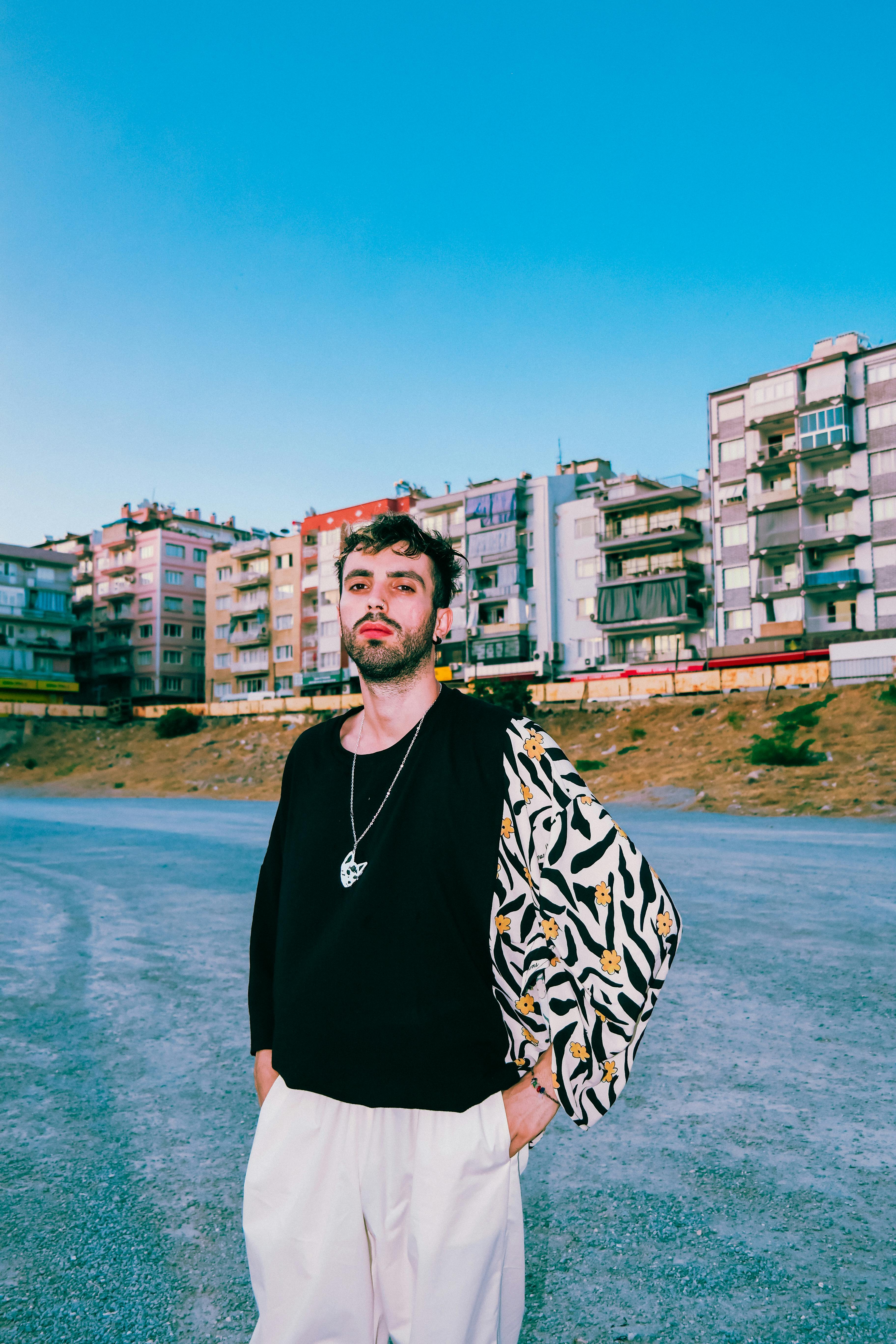A stylish young man poses confidently in front of colorful city buildings under a clear blue sky.