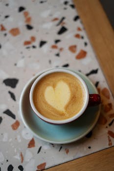 A delicious cappuccino with heart-shaped foam art served in a ceramic cup on wood table.