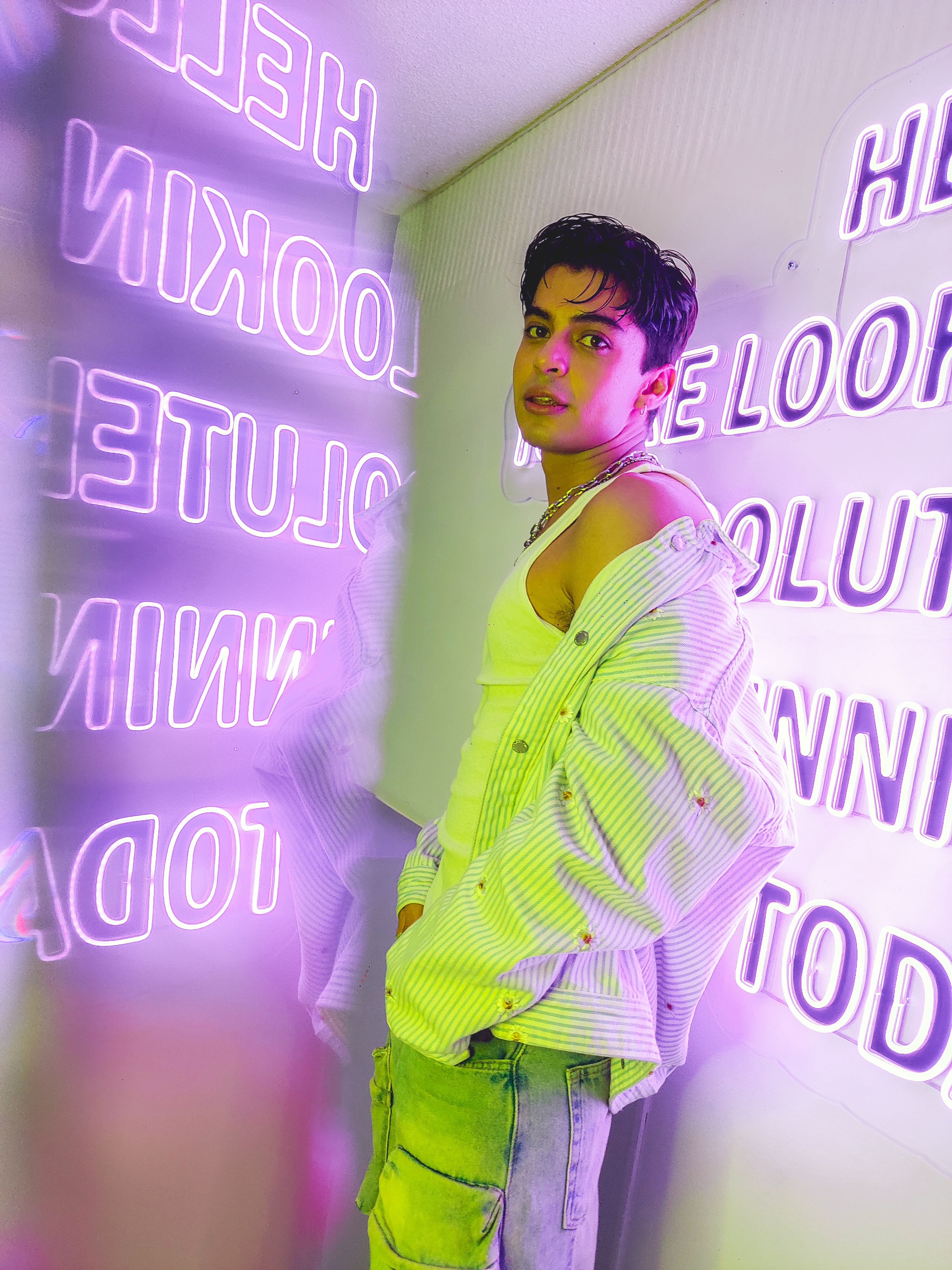 Young man in a stylish outfit posing in a neon-lit room, exuding modern fashion and vibrant nightlife.