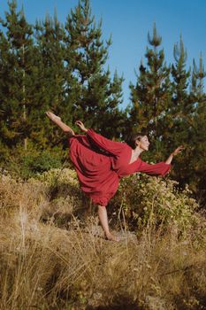 A woman in a red dress elegantly dances amidst a natural setting with pine trees.