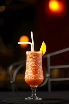A vibrant frozen cocktail served in a glass with an orange slice and two straws against a blurred nightlife background.