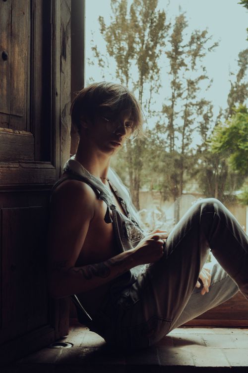 Free Young man in thoughtful pose by a window with soft outdoor lighting. Stock Photo