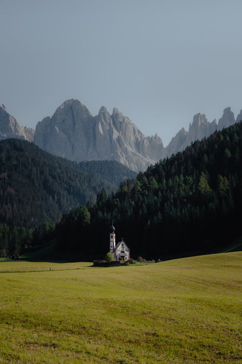 Free A serene chapel nestled in the Dolomites with mountains and a lush green field under a clear sky. Stock Photo