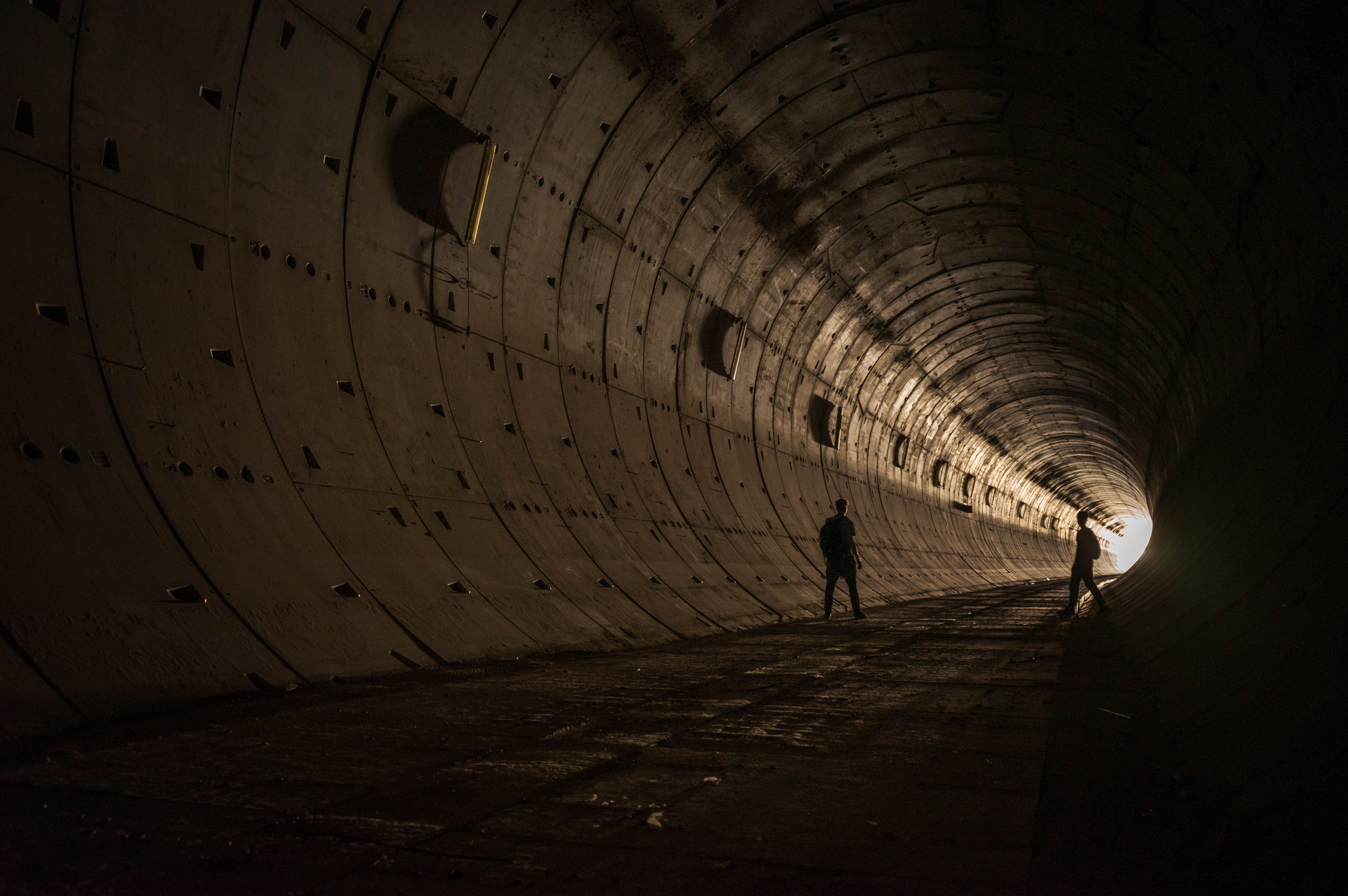 Dramatic silhouettes of two figures standing in an industrial tunnel with light at the end.