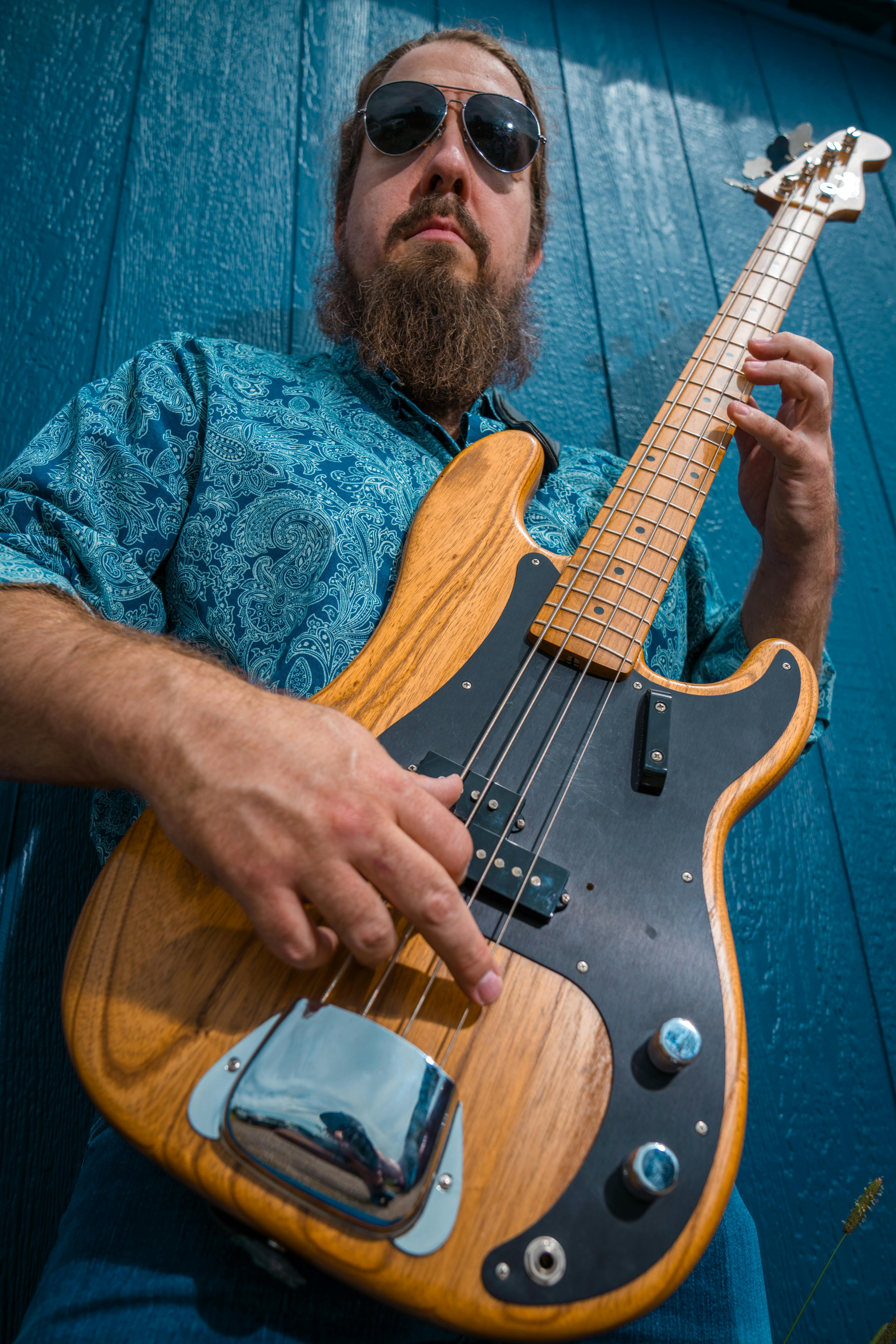 A bearded musician in sunglasses plays a wooden bass guitar outdoors against a blue wall.