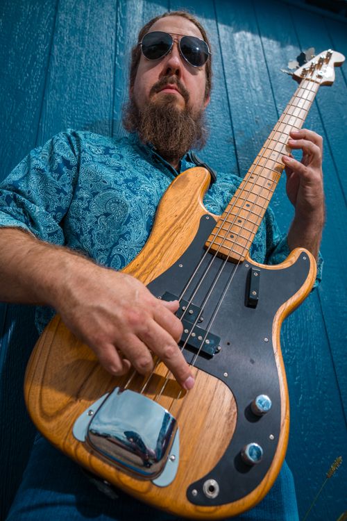 Free A bearded musician in sunglasses plays a wooden bass guitar outdoors against a blue wall. Stock Photo