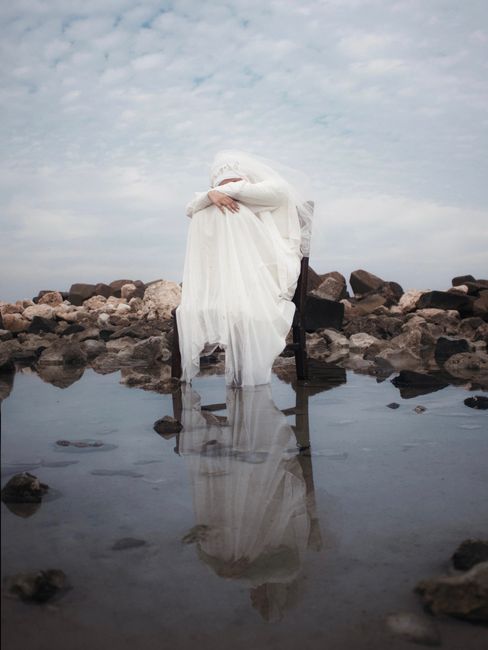 A bride in a wedding dress sits on a chair surrounded by rocks, reflecting in shallow water.