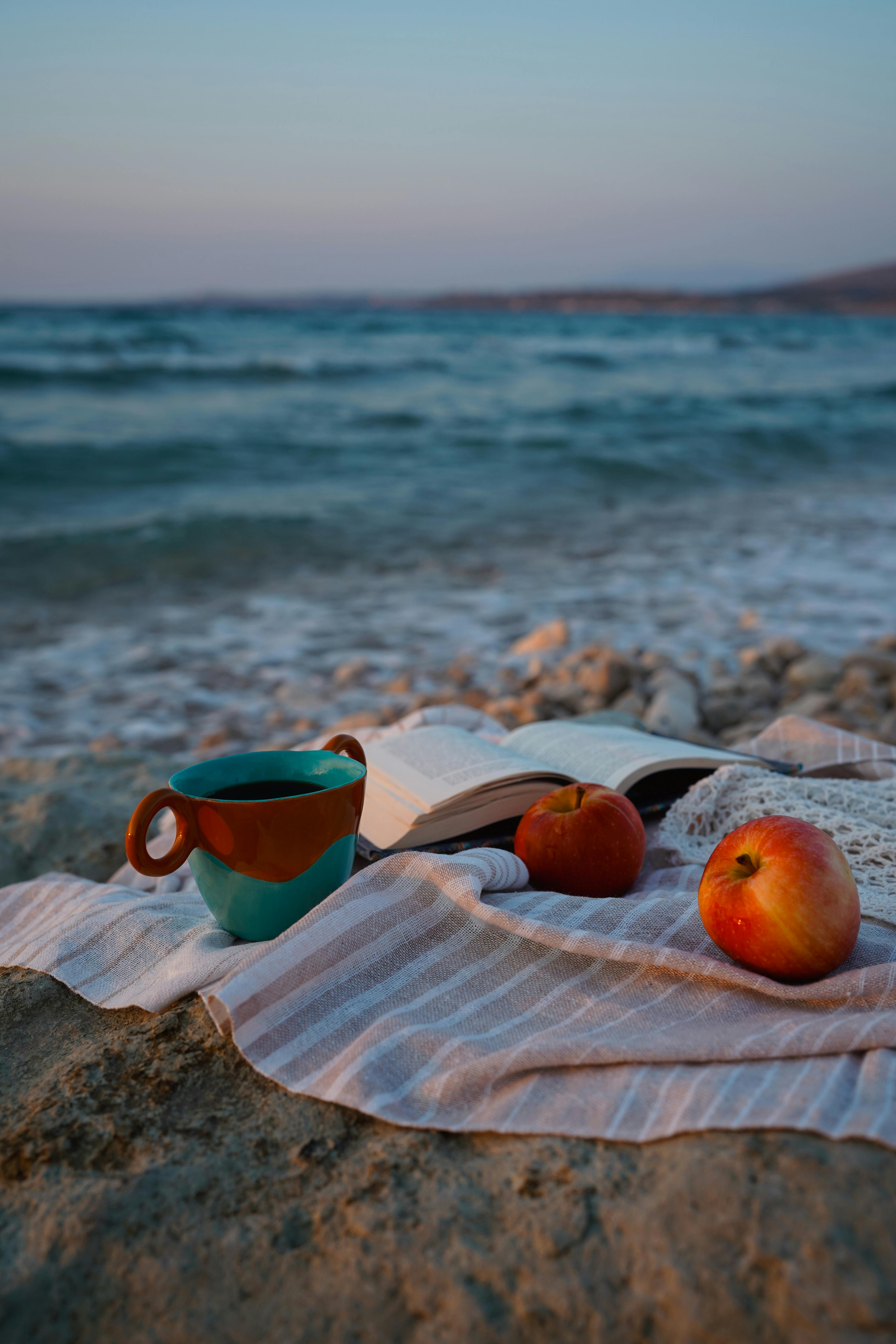 Peaceful beach setup with a mug, open book, and apples during sunset for relaxation.