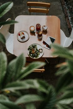 Overhead view of a cafe table with salad, dessert, and drinks, surrounded by green foliage.