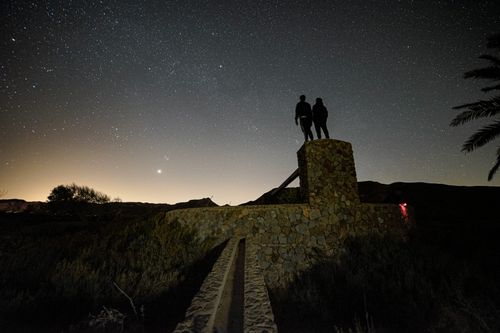 Free Silhouettes of two people on a structure under a starry night sky, creating a dramatic backlit scene. Stock Photo