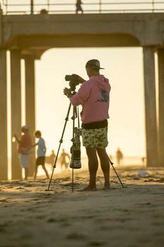 Photographer with tripod capturing a sunset scene on a beach under a pier.