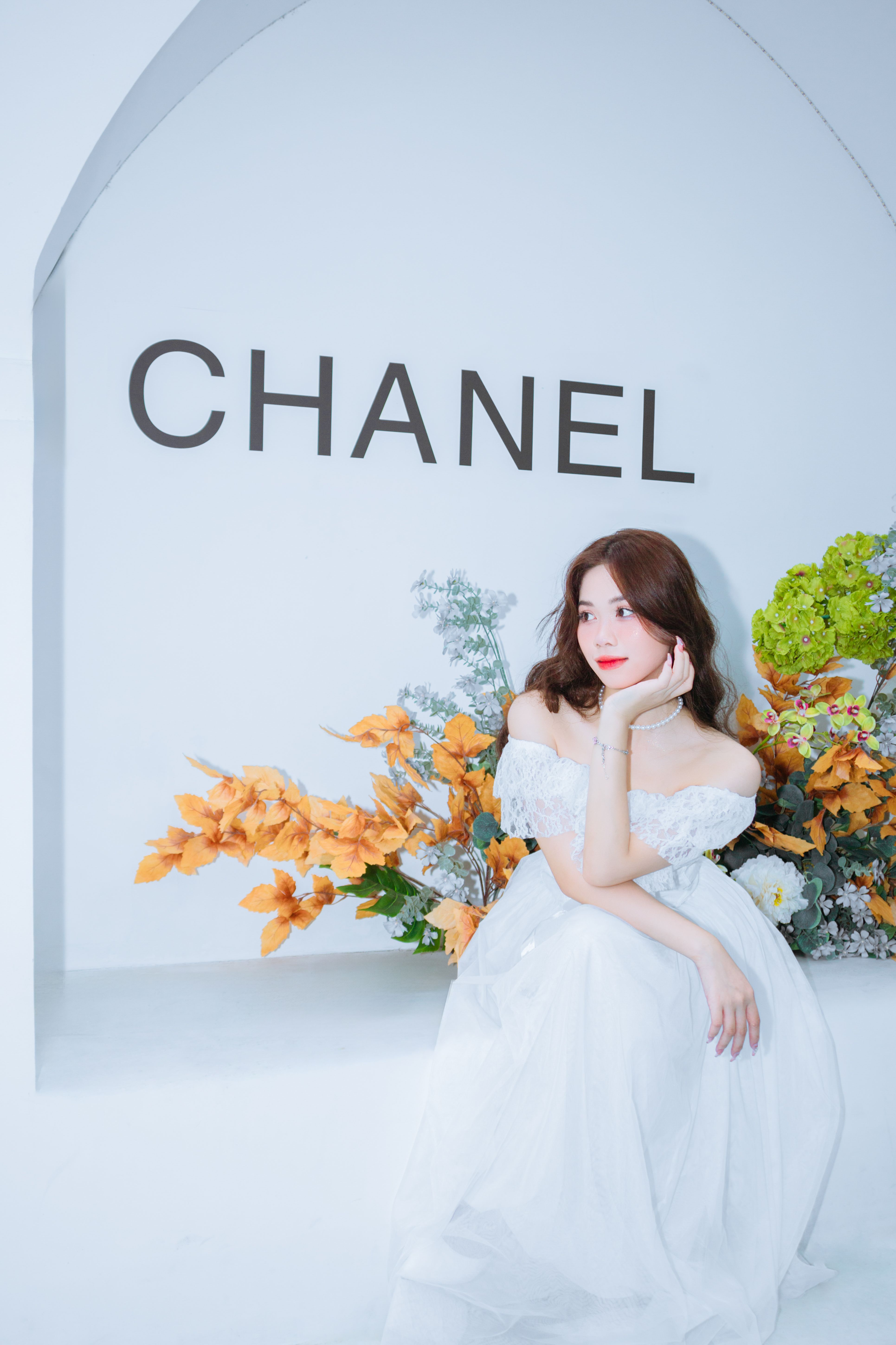 A woman in an elegant bridal gown sits gracefully with vibrant floral decor under the 'CHANEL' sign.