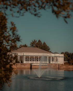 A serene autumn setting with a grand mansion overlooking a tranquil lake and fountain.