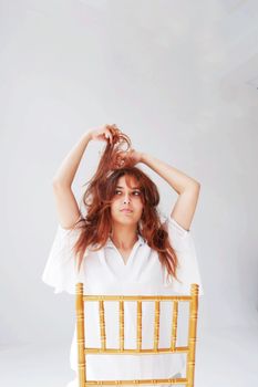 Studio photoshoot of a young woman with red hair posing on a chair, showcasing casual fashion.
