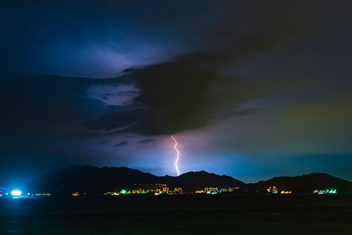 Free Nighttime lightning storm illuminating mountain silhouette with vibrant city lights below. Stock Photo