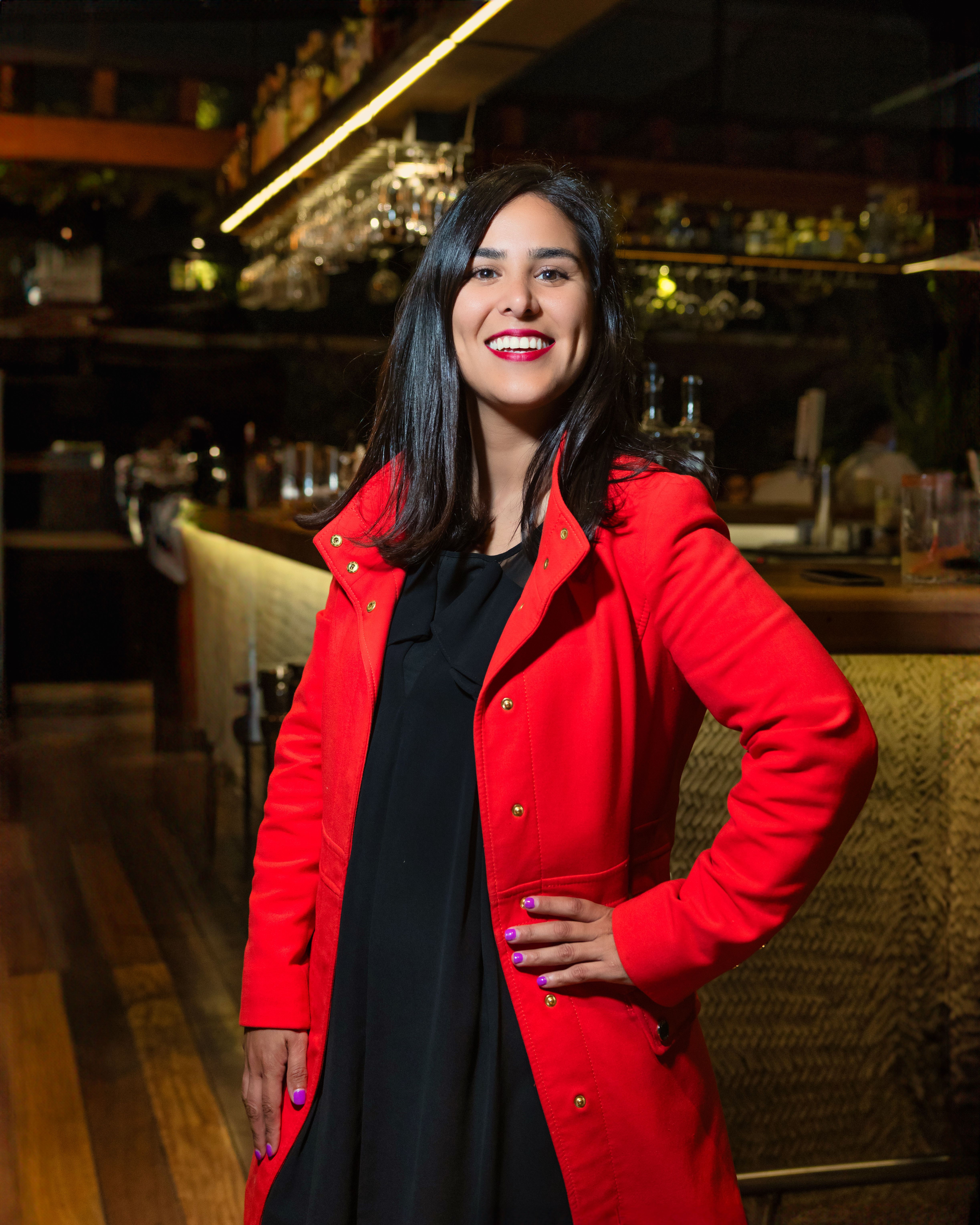 Smiling woman in a vibrant red coat standing confidently at an elegant bar.