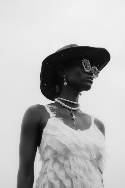 Black and white image of an elegant woman posing in a dress with pearls and a hat.