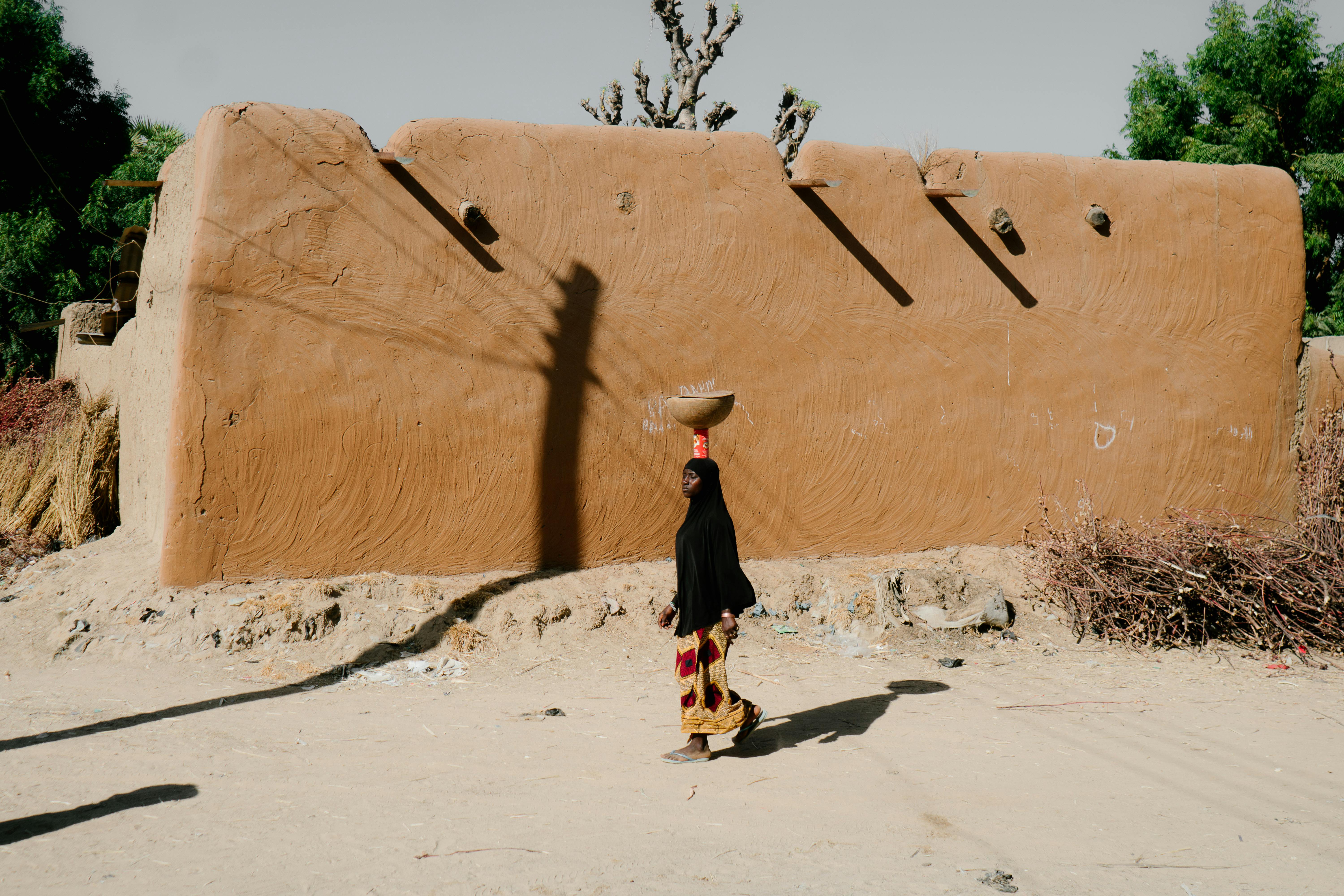 A woman in traditional attire carrying a basket in a desert village setting.