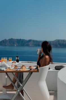 A woman sipping coffee at a table with breakfast overlooking Santorini's stunning ocean view.