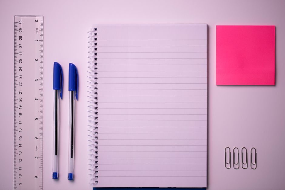 Photo by Nic Wood on Pexels Flat lay of a notepad, blue pens, ruler, sticky note, and paper clips on a pink background.