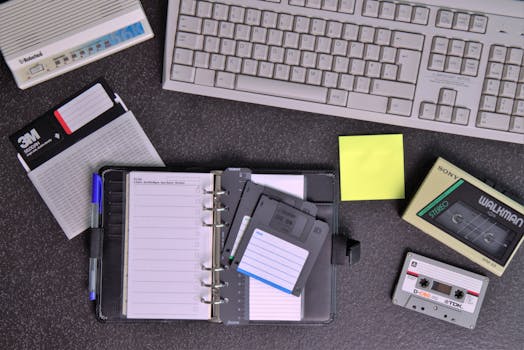Flat lay of retro office supplies featuring a Walkman, floppy disks, keyboard, and planner.