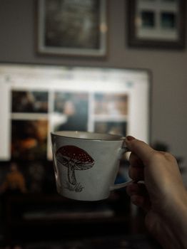 A hand holds a mushroom-themed cup indoors, focusing on coziness with a blurred screen in the background.