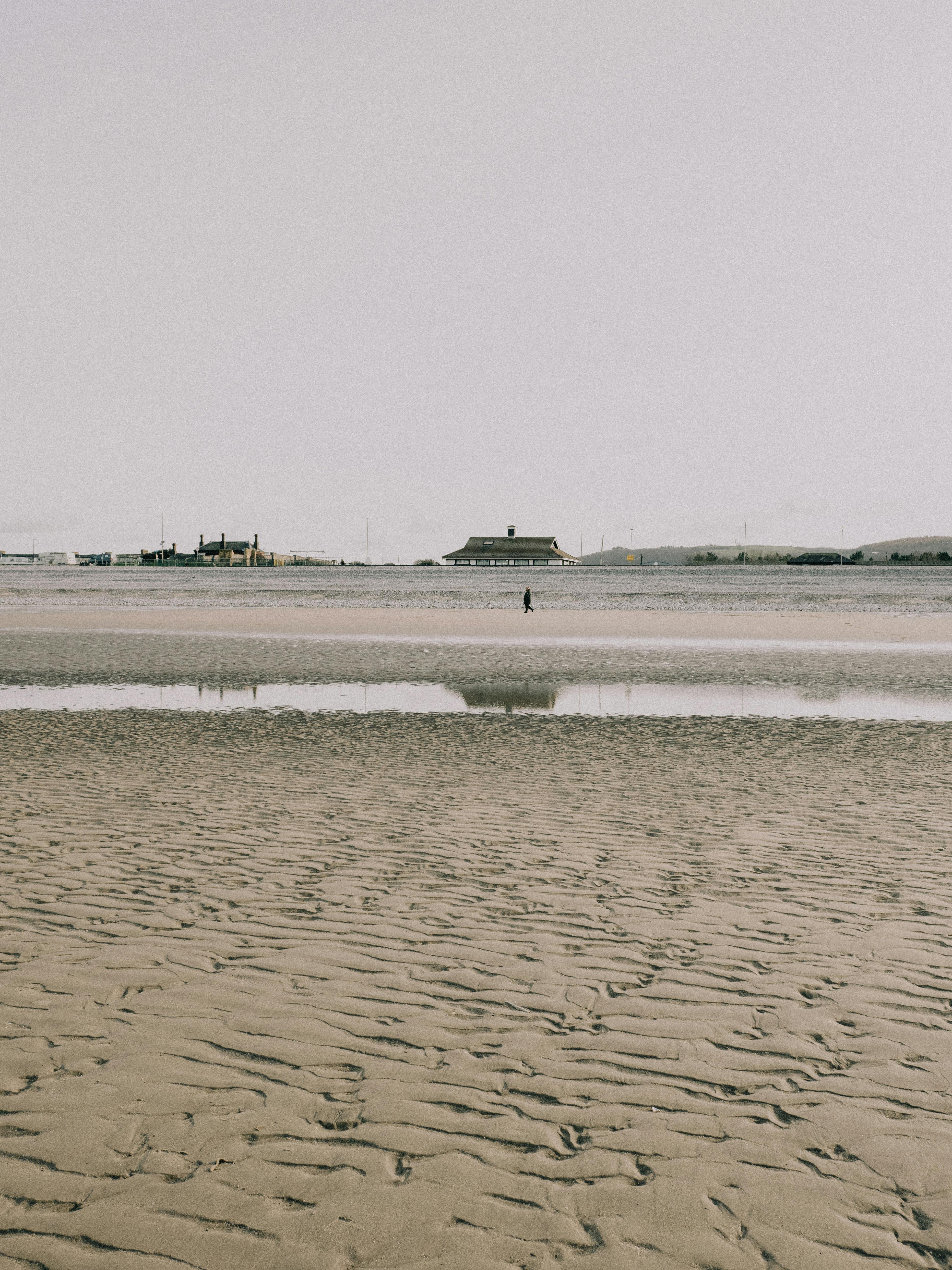 A lone person strolls on a wide sandy beach with distant buildings, creating a peaceful scene.