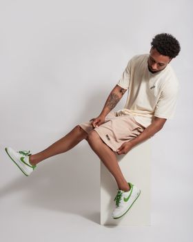 Casually dressed young man with sneakers sitting on a stool in a minimal studio setting.
