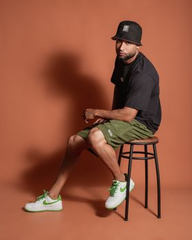Portrait of a young man sitting in casual streetwear on a stool indoors.