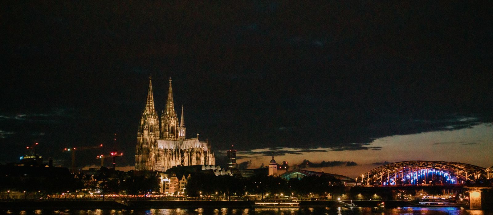 View of Cologne Cathedral and Hohenzollern Bridge illuminated at night, reflecting in the Rhine River — flights from Alicante to Cologne