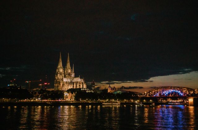 View of Cologne Cathedral and Hohenzollern Bridge illuminated at night, reflecting in the Rhine River — arriving in Köln