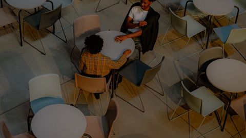Overhead shot of two adults talking at a table in a modern, indoor café with empty tables around.