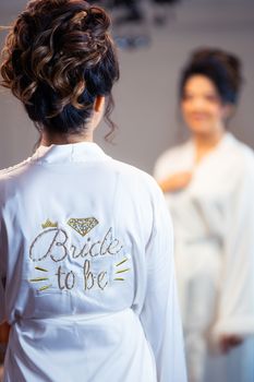 A woman wearing a 'Bride to Be' robe getting ready in the mirror.