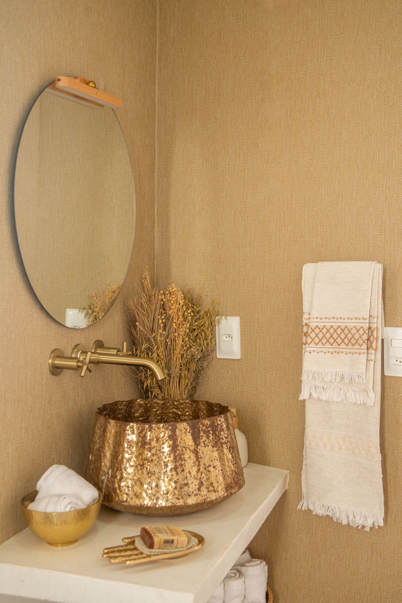 Hammered copper-toned vessel sink with a brass wall faucet, round mirror and a fringed woven towel in a warm southwestern bathroom