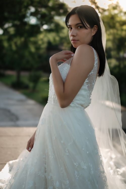 Free A bride poses elegantly in a sunlit outdoor setting, showcasing a modern wedding dress and veil. Stock Photo