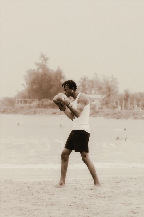 Free A man in boxing gloves practices on a sandy beach, with people swimming in the background. Stock Photo