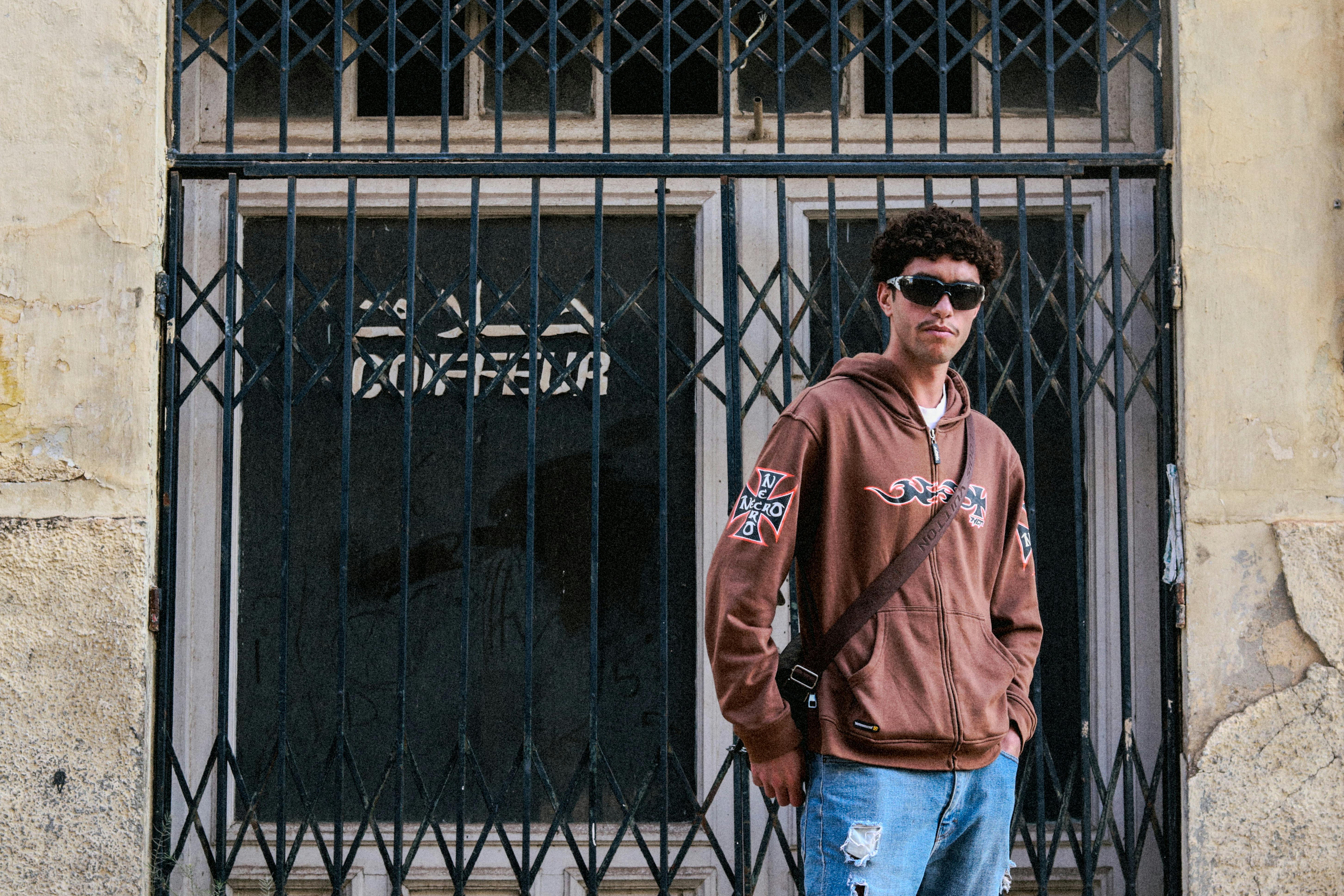 a man in a brown hoodie and jeans standing in front of a metal gate