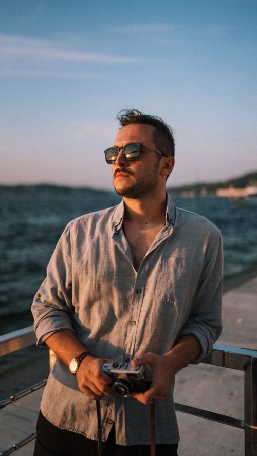 Free Casual portrait of a man in sunglasses holding a camera by the sea during sunset. Stock Photo