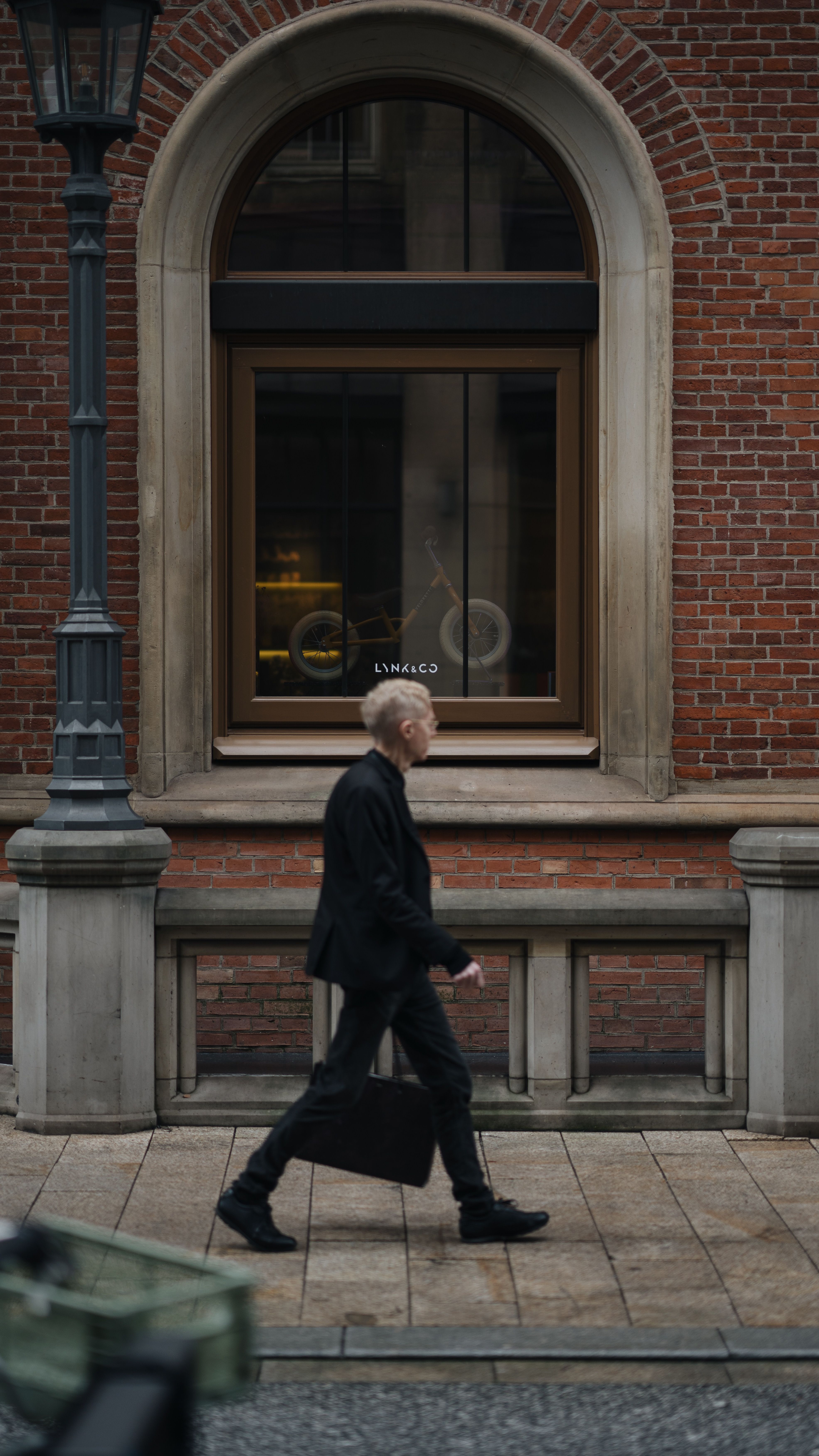 An adult man in business attire walks past a brick building in an urban setting.
